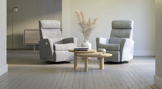 Two light gray upholstered recliners placed on a light wood floor in a bright, minimal room, with a small round wooden coffee table and a vase of dried grass between them.